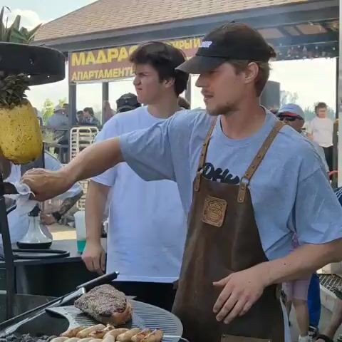 A man wearing a black baseball cap and a brown leather apron is standing to the right of the frame, holding two pineapples on a metal spit above a grill. He is looking to his right. To his left, a large piece of seasoned meat is slowly rotating on a spit over an open fire. Below the rotating meat, another grill holds several rows of sausages. In the background, several people are gathered, watching the cooking process. A building with a sign that says "MADRAS APARTMENTS" is visible behind the crowd. The overall scene appears to be an outdoor event or market.
