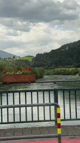 This is a video taken from a moving vehicle, looking out over a bridge. The bridge has a green metal railing. Below the bridge, a wide, greenish-blue river flows. On either side of the river are lush green trees and rolling hills. In the background, more trees cover the hills, with some houses and fields visible on the slopes. The sky is filled with dramatic, grey clouds. The camera is panning to the right, showing more of the landscape as the vehicle moves.