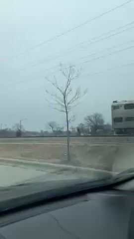 The video shows a Metra commuter train passing on a gray day. The video is filmed from inside a car. The train is silver and blue, and runs parallel to the road. Trees with bare branches are on the side of the road. Power lines run overhead. The weather appears to be wet.