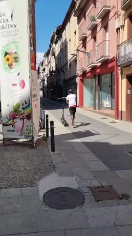 A person on a scooter is traveling down a narrow, European-style street lined with buildings. The buildings have balconies and shops on the ground floor. A large poster is on the left side of the street. The sky is blue and the sun is shining, casting shadows on the street. Another person on a scooter is further down the street and moves towards the first person.