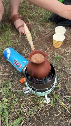The media file shows a close-up of a person making coffee outdoors. The primary subject is a traditional Turkish coffee pot, also known as a cezve, which is made of terracotta and filled with dark coffee. The cezve is placed on a small, portable gas stove, which is fueled by a blue and white gas canister labeled "GAS." The stove has a green base and a black metal heat shield around the cezve. Flames are visible beneath the cezve, indicating it is actively heating.

To the right of the stove, there are two small, disposable paper cups, one on top of the other, with yellow and white patterns. In the background, the legs of at least two people are visible. One person is wearing brown sandals, and the other's hand is reaching towards the cezve, holding a wooden spoon or stirrer. The ground is covered with dry grass and some green vegetation, suggesting an outdoor setting, possibly a park, campsite, or picnic area.

The activity taking place is the preparation of Turkish coffee outdoors. The person's hand is shown stirring the coffee, creating a frothy top. The lighting suggests it might be daytime, though the specific time is not clear. There are no strong indicators of weather conditions, but the dry grass might suggest a dry spell or a season like late summer or early autumn. The overall mood seems relaxed and focused on the process of making coffee. The location context provided is Wazabaa, Abkhazia.