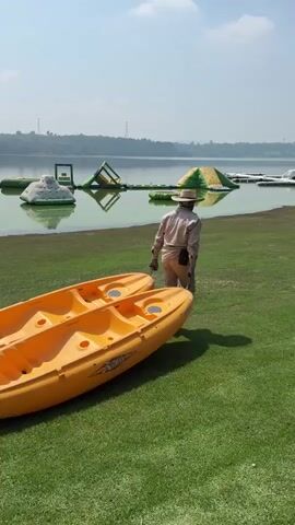 The video shows a person walking on grass near a body of water. In the foreground, there are two orange kayaks. The person, wearing a light-colored hat and outfit, walks away from the kayaks towards the water. In the background, there are various inflatable structures on the water, suggesting a water park or recreational area. The environment appears to be a sunny day with a clear sky and a distant treeline.