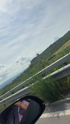 The video captures a dynamic view from inside a moving vehicle, likely a car, in Han Krum, Bulgaria. The perspective is from the passenger side, looking out over a rural landscape.

**Subjects & Scene:** The primary subjects are the natural environment and elements of a highway. A prominent gray guardrail runs parallel to the vehicle's path, separating the road from the adjacent terrain. Beyond the guardrail, a gently sloping hill is covered in vibrant green grass, scattered bushes, and small trees, indicating lush vegetation. In the distant background, a large mountain range stretches across the horizon. The sky is a bright blue with scattered white, puffy clouds, suggesting a clear and pleasant day.

**Objects:** Key objects include the guardrail, green foliage, trees, distant mountains, and clouds. In the immediate foreground, the black casing of the vehicle's side mirror is visible, reflecting a portion of a person's leg or arm, clad in purple clothing.

**Activity:** The vehicle is in motion, presumably traveling along a highway or main road, as evidenced by the passing landscape and the perspective from within.

**Notable Details:** The time of day is clearly daytime, characterized by bright sunlight and visible shadows. The weather appears fair and partly cloudy. The combination of the guardrail, road, and natural hilly/mountainous terrain strongly suggests a drive through a scenic, open area. The location context specifies Han Krum, Bulgaria. No visible text is present in the video. The sounds are consistent with wind noise and possibly vehicle movement, enhancing the feeling of being on the road.