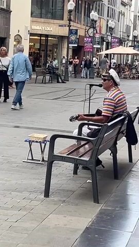 A man is playing guitar and singing into a microphone while sitting on a bench in a public square. He wears a striped shirt, white pants, a white hat, and sunglasses. A black bag sits beside him. A small stool with a basket sits to his left. People walk around in the background. Buildings and streetlights line the square. A storefront for Bimby & Lola is visible. Other people are seated on benches nearby.