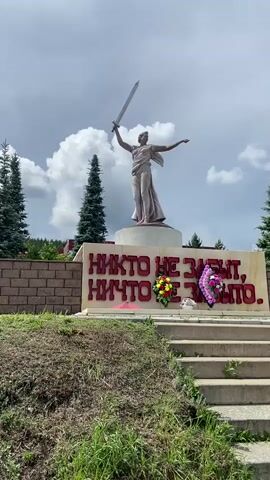 The video shows a statue of a woman holding a sword aloft, positioned on a pedestal in front of a wall with red lettering. The lettering reads "НИКТО НЕ ЗАБЫТ, НИЧТО НЕ ЗАБЫТО" which translates to "No one is forgotten, nothing is forgotten." Two floral wreaths are placed at the base of the wall. The statue and wall are set against a backdrop of evergreen trees, a cloudy sky, and a brick wall to the left. Concrete stairs lead up to the monument from the foreground, which is covered in grass and dirt. The overall scene appears to be a memorial site.
