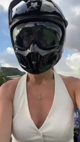 A woman stands outside, wearing a white halter top and a black, angular helmet with goggles. The helmet's mirrored face shield obscures her face initially, but she lifts it to reveal a smiling face. The surroundings suggest a roadside location with buildings and greenery in the background. The sky is overcast.