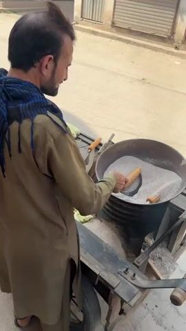 A middle-aged man with a beard is standing behind a black metal stand on a public street. The stand has a single wheel and contains a black drum, which is topped with a large metal bowl filled with a grey grainy substance. The man is wearing a brown tunic and a patterned scarf, and he is inserting corn on the cob into the grainy substance. He uses a metal utensil to move the corn around. Smoke rises from the bowl. The background shows a paved street, buildings with closed metal shutters, and a store. A young boy watches the man from behind the stand.