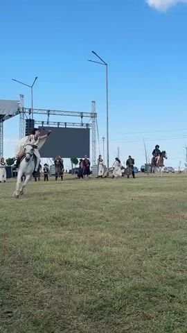 A person dressed in historical costume, wearing a white cape and brown pants, rides a white horse at a gallop across a grassy field. The rider holds a stick or spear in their right hand and their hair is flowing in the wind. In the background, a crowd of people watches the equestrian performance, some standing near tents and vehicles. The sky is blue with a few white clouds. The scene appears to be an outdoor event or festival.