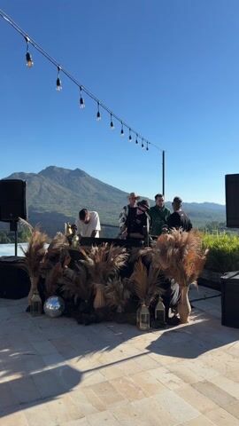 The video shows several people around a DJ setup on an outdoor terrace, with a mountain backdrop under a clear blue sky. String lights hang overhead. The DJ booth is decorated with pampas grass and lanterns. A disco ball sits in front of the booth. There are speakers on either side. Some of the people appear to be setting up, while one DJ waves to the camera.