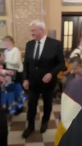 The video shows a man in a black suit handing out gifts to a group of children in what appears to be a museum or an ornate building. The man has white hair and a mustache. He smiles as he hands out small, wrapped items from a blue gift bag adorned with stars. Several children and a young woman wearing a pink jacket surround him, accepting the gifts. The room features patterned tile flooring, decorative walls with artwork and displays, and a doorway in the background. The scene is casual and suggests a holiday or celebratory event.
