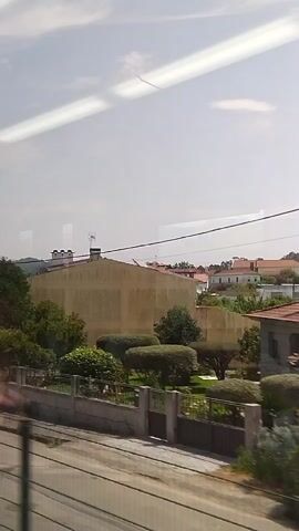 This media file captures a view from a moving vehicle, likely a train, traveling through a suburban or rural area near Barcelos, Portugal. The scene is dominated by a mix of residential buildings and lush greenery.

The subjects include several houses with red-tiled roofs and white or light-colored walls, typical of Portuguese architecture. Some houses are single-story, while others appear to be two-story structures. There are also manicured hedges and trees, suggesting well-maintained properties. Power lines and a utility pole are visible, indicating the presence of infrastructure. The ground level features grass and some dry brush.

The activity taking place is the passage of time and scenery from the perspective of someone traveling. The camera is likely positioned inside the vehicle, looking outwards through a window, as indicated by the streaks of light and reflections on the glass.

Notable details include the bright, overcast sky, suggesting it is daytime, likely late morning or afternoon. The overall impression is one of a peaceful, sunny day in a developed area surrounded by nature. The reflections on the window create a slightly hazy or ethereal quality to the scene. The location cues, such as the architectural style and the mention of Barcelos, Portugal, place the scene specifically in that region. There are no visible text elements in the media file.