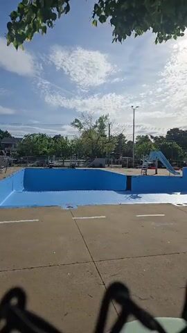 The video captures an outdoor public swimming pool area in Laval, Canada, under a clear, sunny sky with scattered white clouds. The swimming pool itself is empty or in the process of being filled or drained, with a small amount of blue water visible at the very bottom of the blue-painted basin. The surrounding area consists of a concrete deck, which appears dry.

Key features of the pool facility include a light blue water slide visible in the background, positioned to enter the pool. On the right side of the frame, a metal ladder with handrails provides access into the empty pool. Further in the background, additional pool equipment and structures are partially visible. The entire area is bordered by green trees, with their leafy branches framing the top of the video shot. A tall light pole stands in the mid-ground. The footage appears to be recorded from just outside the pool enclosure, with the top of a chain-link fence visible at the bottom of the frame.

The scene is devoid of people, suggesting the pool is not in active use, possibly due to it being off-season or before opening hours. The time of day appears to be daytime, likely morning or early afternoon, given the bright sunlight and shadows cast by the ladder. No visible text is discernible in the video. The atmosphere is quiet and serene, anticipating the warmer days when the pool will be filled and in use.