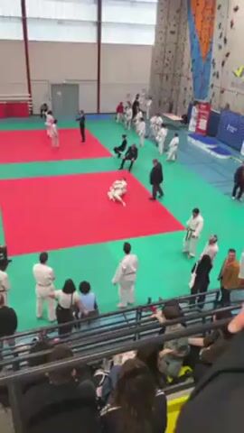 The video shows a judo match taking place on a red mat. Two individuals in white judogi uniforms are grappling on the mat. One of the judokas wears an orange belt. Several spectators watch the match from the sidelines and the stands. Some individuals are wearing judogi uniforms while others are in casual clothing. Referees dressed in suits are present to oversee the match. The event appears to be taking place indoors in a gymnasium, indicated by the mats, wall-mounted climbing equipment, and high ceiling.