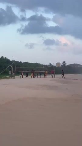 This media file depicts a group of young people playing volleyball on a sandy beach. The scene is set during what appears to be late afternoon or early evening, with a soft, diffused light and a sky filled with dramatic, billowy clouds, suggesting possibly overcast or approaching weather.

The subjects are a mix of individuals, predominantly appearing to be male, dressed in casual beachwear. They are actively engaged in a game of volleyball, with some players positioned on one side of the net and others on the opposite side. The volleyball net is clearly visible, strung between two poles. The sand is a light, sandy color, and the foreground is dominated by the beach.

In the background, there is a line of lush green vegetation, possibly trees and bushes, characteristic of a coastal environment. Beyond the greenery, structures are visible, including what looks like a building with a darker facade and a tall, slender tower. Palm trees are also scattered in the background, further reinforcing a tropical or subtropical location. The overall atmosphere seems relaxed and recreational, with the focus on the group enjoying the game. There is no visible text in the media file.