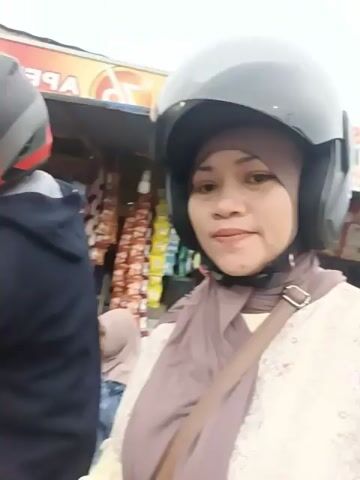 A person wearing a helmet and a hijab is filming a video, turning the camera to show another person, also wearing a helmet, who is holding a snack. The background appears to be a street stall or market, with various items displayed for sale, including snacks and drinks. Two children, one boy and one girl, are also visible in the background, seemingly interacting with the people at the stall. The overall setting suggests a casual outing or a stop at a local vendor.