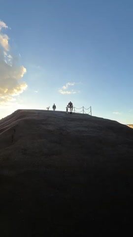 A medium shot captures a man and a young child descending a small, man-made hill against a backdrop of a bright, clear blue sky with scattered clouds. The hill is made of a dark, granular material, likely asphalt or compacted dirt. A chain fence, supported by wooden posts, runs along the edge of the hill. The man, wearing shorts and a t-shirt, is carefully guiding the child, who is wearing a matching outfit. Another person stands on the hill, taking a photo, and there is an animal further in the background. The sun is setting, creating a soft, golden light on the horizon. The overall impression is one of a peaceful, family-oriented moment.