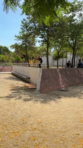 A person on a skateboard is performing a trick on a rail in an outdoor skatepark. The skateboarder is wearing a black t-shirt and black pants, with tattoos visible on their arms. The skatepark has a concrete wall with a brick-like texture on the lower half and a white wall on the upper half. There are trees and a clear blue sky in the background. Other people are sitting and watching the skateboarder. The ground is covered in dirt and fallen leaves.