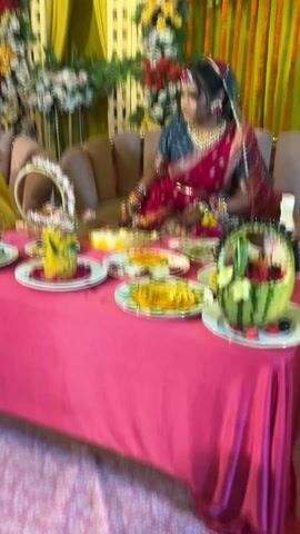 A wide shot depicts a seated woman in traditional dress, likely a bride, behind a table laden with fruit platters. She wears a maroon and teal sari and heavy gold jewelry. The table is covered with a pink tablecloth. Behind her is a decorative backdrop with flowers and yellow fabric panels. The camera tilts upwards, revealing an elaborate ceiling decoration with colorful fabrics and string lights. As the camera zooms out, a second woman in a pink hijab is seen arranging fruit on the table. A young child in a red and yellow outfit is also present. The scene suggests a wedding or other celebratory event.