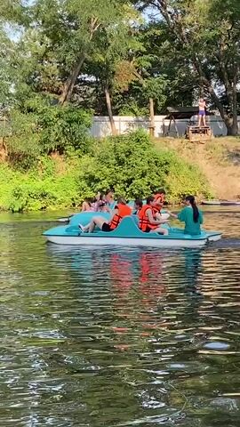 The video shows a turquoise paddle boat with five people on a body of water. Three individuals are in the front half of the boat, two of whom appear to be pedaling, and all are wearing orange life vests. Two individuals are in the back half of the boat, one in a green shirt. The boat is moving from left to right. In the background, there is lush green foliage and trees, and a light-colored fence along the top of a grassy embankment. Another paddle boat is visible in the distance to the right. The water is green and reflective, showing the sky and trees.