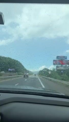 The video is filmed from the interior of a vehicle, looking out through a windshield covered in raindrops. The view shows a highway with several cars driving ahead in the same direction. The sky is cloudy with patches of blue visible. Green hills covered in vegetation are on either side of the highway. A large billboard is visible on the right side of the road. The car appears to be moving at highway speed, as indicated by the motion blur of the passing scenery and the raindrops on the windshield.