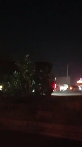 At night, the camera, possibly in a moving vehicle, captures a scene of a busy road. Multiple vehicles, including cars and trucks, are visible with their headlights illuminated. The road is bordered by trees and buildings, some of which have signs. A large advertising board is also present, but its specific content is unclear due to the low light and distance. The overall atmosphere is typical of a nighttime urban or suburban environment with moderate traffic.