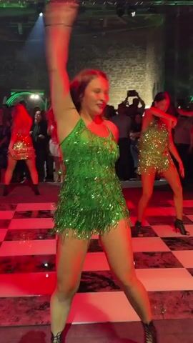 The video shows a group of people dancing on a checkered dance floor in a dimly lit room. There are spotlights shining on the dancers. Several women are wearing short, sequined dresses and dancing. In the background, there are people standing and watching the dancers. The room appears to be a venue with stone walls and a ceiling with visible lighting fixtures.
