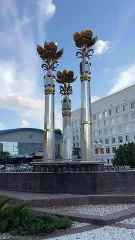 A large, ornate fountain with three metallic columns topped with golden flower-like structures stands in the foreground. Water sprays from the top of the central column and cascades down the sides of the fountain's basin. The fountain is surrounded by a dark, tiled base, and in front of it, there are manicured green bushes and white gravel. In the background, a modern white building with many windows rises up. To the left of the fountain, a smaller building with a blue roof and large glass windows is visible, reflecting the sky. The sky is blue with scattered white clouds. The overall scene is an outdoor public space, likely a plaza or square. No people are clearly visible in the immediate vicinity of the fountain, though there might be some distant figures near the background buildings. The lighting suggests it is daytime, possibly late afternoon given the angle of the sun.
