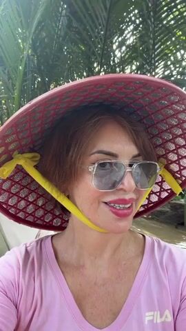 A medium close-up video shows a smiling woman posing for the camera, likely taking a selfie or being filmed. She has reddish-brown hair styled in a short bob with bangs and is wearing large, clear-framed sunglasses. Her lips are painted with bright red lipstick. She wears a pink short-sleeved V-neck shirt, on which the brand name "FILA" is faintly visible in white on the lower right side.

The most prominent object is a traditional Vietnamese conical hat (nón lá) made of red woven material, with a yellow chin strap tied in bows at the sides. The hat is intricate, with a patterned, possibly plastic or bamboo, lattice design.

The background is somewhat blurred but appears to be outdoors, featuring lush green foliage, consistent with a tropical environment. The lighting is bright and natural, suggesting it is daytime. The woman has a happy and relaxed expression. Based on the provided context, the scene is set in Thoi Son, Vietnam.