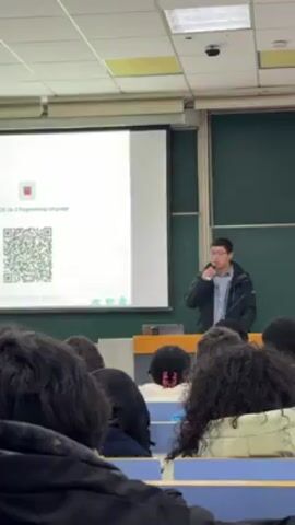 A man in a dark jacket and glasses stands behind a wooden podium, addressing a classroom full of students. He holds a microphone to his mouth. Behind him, a projector displays a slide labeled "25-06-2 Programming Language" along with a QR code. The students sit at desks, their backs and heads visible in the foreground. The classroom has a green chalkboard and pale green walls. The scene appears to be a lecture or presentation in a university setting.