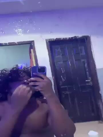 A shirtless person with dark skin and shoulder-length dark curly hair styles their hair in front of a mirror while holding a blue smartphone to take a selfie. The background shows a white wall with two doors, one with a greenish-blue tint and one with a dark brown peeled finish. The room has a messy appearance, with possible water damage or peeling paint on the wall near the mirror, which also has some glittering dust-like particles on its surface. The lighting changes color during the video, alternating between a normal white light, a red hue, and a green hue.