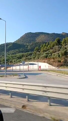 The video shows a view from inside a moving vehicle on a sunny day. The landscape is hilly and covered with green vegetation. In the foreground, a highway guardrail is visible. Beyond the guardrail, there are several buildings, including what appears to be a gas station with fuel pumps and a convenience store. There are also covered parking areas. Signs for "EKO" and "BP" are visible at the gas station. The sky is clear and blue. The video appears to be a short clip, likely from a dashcam or a handheld camera mounted inside the vehicle.