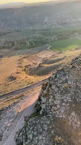 The video captures a panoramic view of a vast, rugged landscape at either sunrise or sunset. The camera initially points down a steep, rocky slope, revealing a valley below with green agricultural fields, a winding dirt road, and scattered buildings. The sun is low on the horizon, casting a warm, golden light across the scene.

As the camera slowly pans to the right, it reveals undulating hills, distant mountains, and a clear sky that transitions from bright blue overhead to soft orange and yellow near the horizon. A body of water, possibly a lake or river, is visible in the middle distance. The pan concludes with the sight of at least two hot air balloons rising gracefully against the backdrop of the sunrise or sunset, adding a picturesque element to the already scenic view. The terrain is largely dry and arid, with sparse vegetation, contrasting with the vibrant green patches in the valley.

The sounds of a person speaking in Russian, commenting on the landscape (possibly comparing it to the moon), and laughter are heard, suggesting an appreciative and joyful atmosphere among unseen observers. The setting is Yeşilhisar, Turkey.