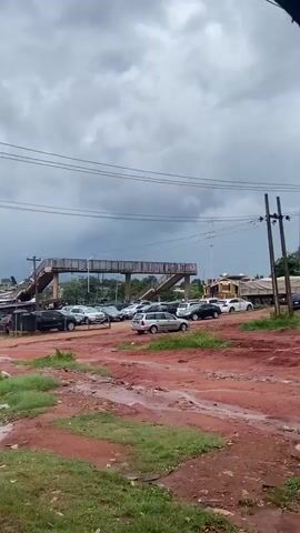 The image depicts an outdoor scene in what appears to be a semi-urban or suburban area in Auchi, Nigeria, under an overcast sky. The foreground is characterized by a sprawling, reddish-brown dirt expanse, deeply rutted and muddy in sections, indicative of recent rain or poor road conditions. Sparse patches of green grass are visible amidst the dirt.

In the mid-ground, a silver station wagon, positioned with its rear facing the viewer, is parked on the dirt. Behind it, numerous other cars of various models, including sedans and SUVs in shades of silver, black, and white, are parked seemingly haphazardly across the unpaved lot.

Dominating the background is a sturdy pedestrian bridge, likely constructed from concrete or metal, with railings along its sides. Several people are visible walking across the bridge, with others ascending the stairs on its right side. Above the bridge, multiple power lines crisscross the sky. To the far right in the background, a building with a light-colored roof and possibly yellow walls is partially visible. The sky is heavily clouded with dark grey clouds, suggesting an overcast day. No discernible text is present in the image.
