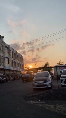 A vertical video shows a street scene during sunset. In the foreground, a dark asphalt road is visible with some debris and a small mound of dark material. To the right, a silver car is parked, facing away from the camera, with its hood and roof appearing to be black or a darker shade. Further to the right, another vehicle is partially visible. In the midground, to the left, a multi-story building with several floors is shown. The building has a facade with alternating colors and numerous windows. Along the sidewalk in front of the building, several people are gathered, and there are motorcycles parked. A dark-colored pickup truck with a load in its bed is positioned in the street between the building and the parked silver car. The sun is setting behind a line of trees, casting a warm glow and visible rays. The sky above is a mix of pale blue and orange hues with scattered clouds. Power lines are visible crisscrossing the upper portion of the frame. In the background, a large vessel, possibly a ferry, with multiple decks and railings, is visible near the water's edge. The overall scene depicts a busy street or market area by the coast as day turns into evening.