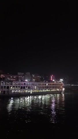 The video shows a body of water, probably a river, with multiple large, illuminated party boats or houseboats on it at night. The water reflects the lights from the boats, creating shimmering patterns. In the distance, there are some buildings with lights, indicating an urban area. The sky is dark and clear. Some people can be seen on the closest boat.