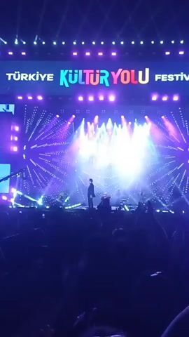 The video shows a concert stage from the perspective of someone in the audience. The stage backdrop reads "Türkiye Kültür Yolu Festivali" in large letters, lit up with purple and white lights. A performer, silhouetted in a dark suit, stands near the center of the stage in front of a microphone. Other musicians and equipment are visible, though less distinct. The stage is illuminated with a dynamic display of bright spotlights and laser-like effects. Below the stage, the audience can be seen holding up phones to record the performance. The atmosphere is dark except for the stage lighting.