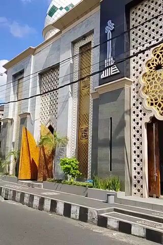 A drone view pans across the facade of a mosque. The mosque has a gray stone exterior with ornate gold-colored geometric patterns above the entrance. Arabic calligraphy is visible on the facade. The mosque has a dome and a tall, thin minaret. The sky is blue with some clouds.
