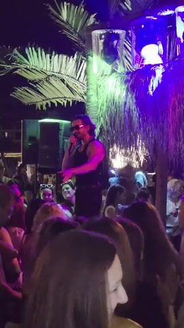 A man with long brown hair and sunglasses stands on a raised platform, singing into a microphone. He is wearing a black tank top and black pants with a belt. The platform is decorated with palm fronds. Behind him, there are purple and green lights. In front of the platform, a crowd of people, mostly women, are watching him. The scene appears to be outdoors at night.