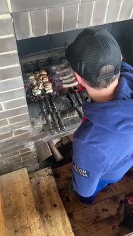 A person wearing a blue hoodie and a black baseball cap is grilling meat on skewers over a fire in a brick fireplace. The person is using tongs to adjust the meat. There are several skewers with meat on them, and some of the meat appears to be shish kebab. The fireplace is made of red brick, and there are wooden steps leading up to it. The floor is made of wooden planks. The person is looking down at the meat, and their face is partially obscured by their hat. The video is from a high angle, looking down at the scene.