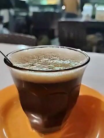 A glass of coffee sits on an orange saucer on a white table. A spoon is in the glass. The coffee has a layer of foam on top. In the blurred background, a person without a shirt walks behind a dark counter, and there are lights hanging.