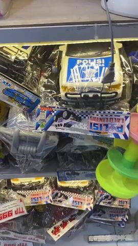 This is a close-up, handheld video that pans across shelves displaying various toys and merchandise. The scene appears to be a retail environment, possibly a toy store or a market stall.

At the beginning of the video, a white toy police car with blue markings is visible, packaged in plastic. Next to it, a colorful stacking toy with cartoon animal features is also displayed. The camera then pans to the right, revealing rows of packaged items, many of which appear to be trading cards or small toy collectibles. These packages feature popular characters and brands, including "Free Fire" and "Spider-Man".

The lighting suggests an indoor setting, likely with artificial lights illuminating the merchandise. The overall impression is one of a display of toys and collectibles, possibly aimed at children or collectors. There are no people clearly visible in the frame, and the focus is solely on the products being showcased. The video is brief, with the camera movement being smooth but not entirely steady, indicating it was likely recorded by someone browsing the items.