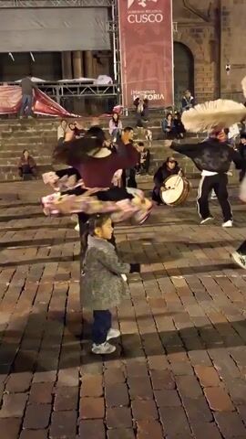 A group of people are performing a traditional dance on a brick-paved area. A woman in a maroon top and a colorful skirt is dancing with a little girl in a gray coat. A man plays the flute while another man plays a large drum. Behind them, people are sitting on steps watching the performance. A banner in the background reads "¡Hagamos Historia!" The setting appears to be an outdoor public space. The lighting suggests it's either dusk or evening.