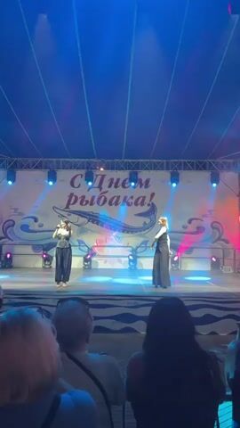 This is a vertical video of a stage performance. Two women are singing and dancing on stage. They are both wearing black outfits. The stage is lit with blue and pink lights. Behind them, there is a banner that says "С Днем рыбака!" which translates to "Happy Fisherman's Day!". There is a large image of a fish on the banner. In the foreground, the backs of audience members' heads are visible. The audience appears to be watching the performance. The overall environment is an outdoor venue, possibly a festival or concert.