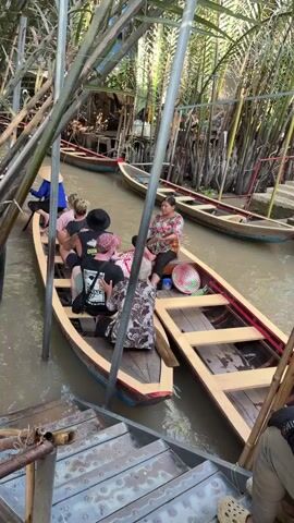 A group of people, appearing to be tourists, are seated in a long, narrow wooden boat on a calm waterway. One person at the front of the boat is wearing a blue conical hat and is paddling. Another individual at the back of the boat also appears to be paddling with an oar. A woman in a floral shirt is seated in a separate, similar wooden boat alongside, holding an oar and observing. Both boats have distinctive painted red and blue stripes on their exteriors. Lush green tropical vegetation, possibly palm trees or similar foliage, lines the banks of the narrow waterway. Wooden poles or structures are visible in the water, suggesting a dock or mooring area. The scene depicts a boat tour or local transportation through a canal, likely in a rural or semi-rural setting during bright daylight. The conical hats and the type of boats are characteristic of the Mekong Delta region in Vietnam.