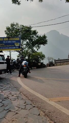 A motorcyclist wearing full riding gear is slowly pulling away from a parking spot on a paved road. The motorcycle is black and has luggage strapped to the back. The motorcyclist is surrounded by other parked motorcycles and a white SUV, beside which stands a person in black attire. To the left of the vehicles is a blue sign, advertising "ANH ANH MASSAGE" in Vietnamese, and the text "XE ĐƯA ĐÓN MIỄN PHÍ" which translates to "FREE PICK-UP". In the background, a large mountain looms under a hazy sky. The road follows the curve of the landscape, with a low metal guardrail separating it from a steep drop-off. Patches of trees and vegetation are scattered around the area.