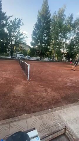 The video shows a tennis court with a clay surface. In the foreground, there is a bench with several items on it, including a red backpack, a blue cap, a water bottle, and a tennis racket. The camera is positioned to the side of the court, looking towards the net and the opposite side of the court. There are trees and buildings in the background. A person is visible on the tennis court playing tennis. The person is wearing dark clothing and is hitting a tennis ball with a racket. The sun is shining, and the sky is blue. The video is shaky, suggesting it was recorded handheld.