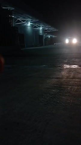 The video captures a dark scene outside what appears to be a warehouse or industrial building. The building has a row of covered loading docks with bright lights illuminating the area. In front of the building, a paved surface, possibly asphalt, is visible with puddles or wet patches reflecting the light. Several vehicles with their headlights on are driving towards the viewer or parked in the distance, creating bright flares in the darkness. The overall atmosphere is dim and hazy, suggesting it might be foggy or misty. The camera is positioned low to the ground, looking slightly upwards towards the building and the vehicles.