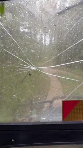 The video shows the view from inside a vehicle, looking through a cracked windshield. It is raining, and raindrops are visible on the glass. The vehicle appears to be moving on a dirt path through a wooded area. The path curves to the right. On the right side of the windshield, a red and yellow reflective marker is visible. The interior of the vehicle is partially visible at the bottom of the frame, showing part of a dashboard. The video also briefly shows a side view through the windshield, revealing the rear wheel of the vehicle splashing through mud and vegetation. The overall impression is of a rough drive through a natural environment in wet weather.