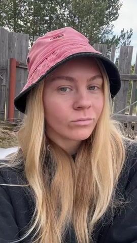 A woman with blonde hair is centered in the frame, wearing a pink and black bucket hat and a black sweatshirt. She is looking directly at the camera with a neutral expression. Her hair is long and straight, falling over her shoulders. She briefly tucks some hair behind her ear with her right hand.  Behind her, a weathered wooden fence is visible, along with some greenery and trees. The sky is overcast. The shot appears to be taken outdoors during the day.