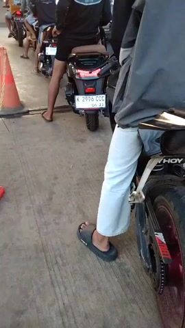 The video shows a line of motorcycles at what appears to be a gas station. The camera is positioned behind a motorcycle in the foreground, focusing on the back of the rider's head and shoulders. The rider is wearing a black and gray jacket and white pants. Their feet are bare, and they are wearing black slide sandals. The motorcycle is black and has a red taillight. In the middle ground, another motorcycle is visible, with a rider wearing a black long-sleeved shirt and black shorts. This motorcycle has a license plate that reads "K 2996 CCC". Further back, other motorcycles and riders are visible, forming a queue. Orange traffic cones are placed along the side of the road. The ground is paved with concrete. The sun is setting in the background, casting a warm glow. The overall scene suggests a typical day at a gas station, with people waiting to refuel their vehicles.